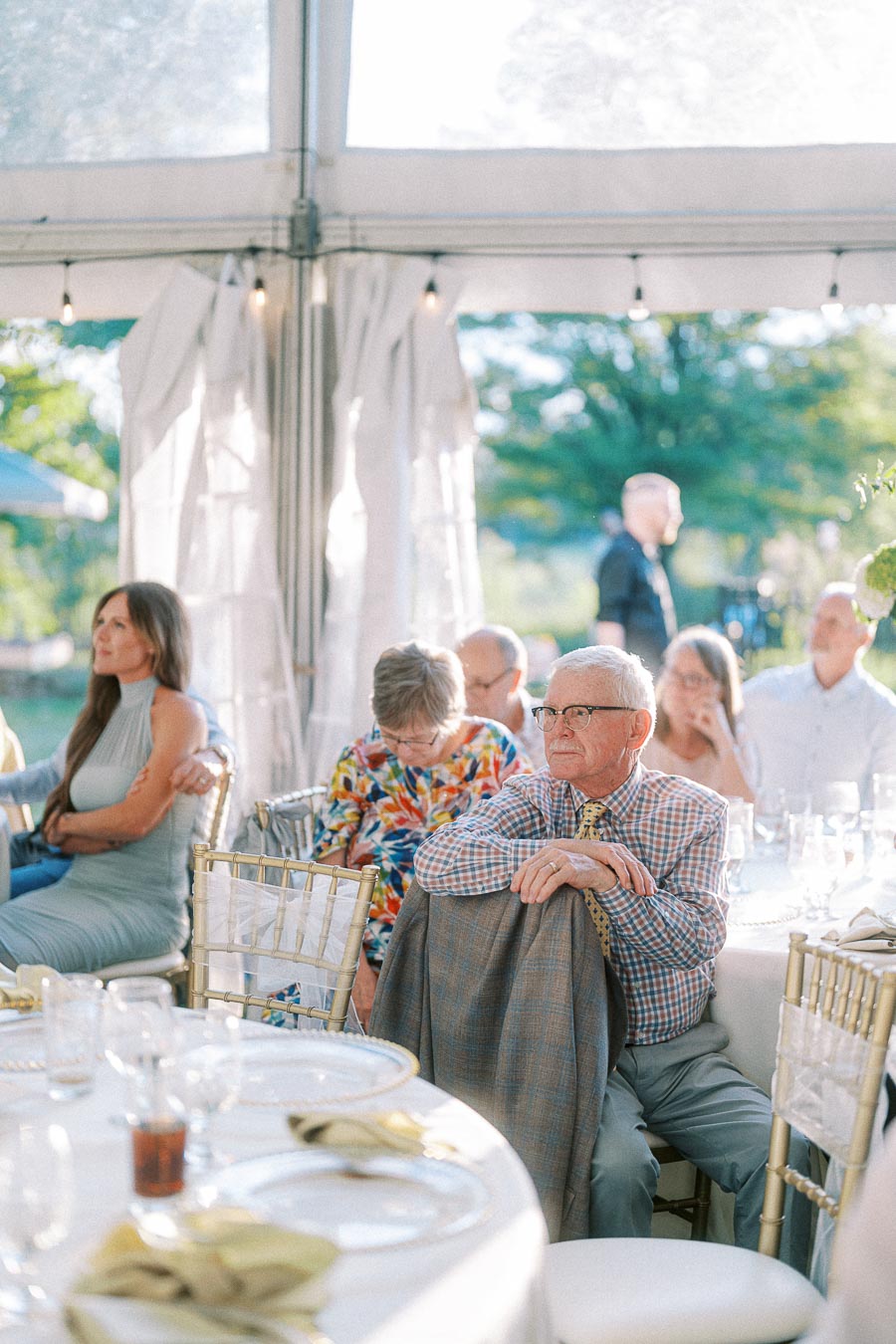 Elderly man sitting at a wedding banquet table under a tent, surrounded by guests, wearing plaid shirt and tie, thoughtful expression, natural outdoor lighting.