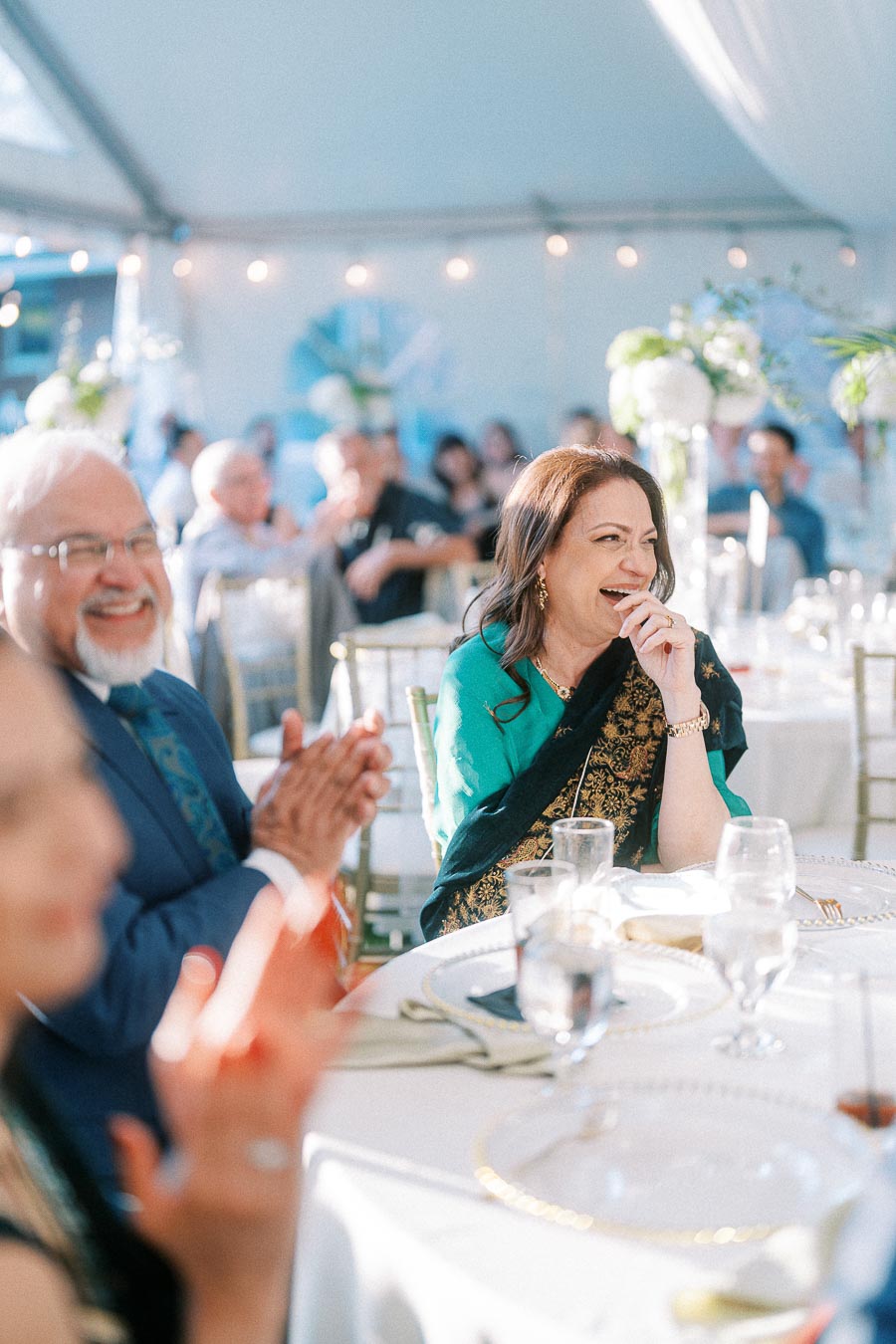 A joyful woman in a colorful saree laughs and gestures while seated at a festively decorated table during an indoor celebration, surrounded by other smiling guests.
