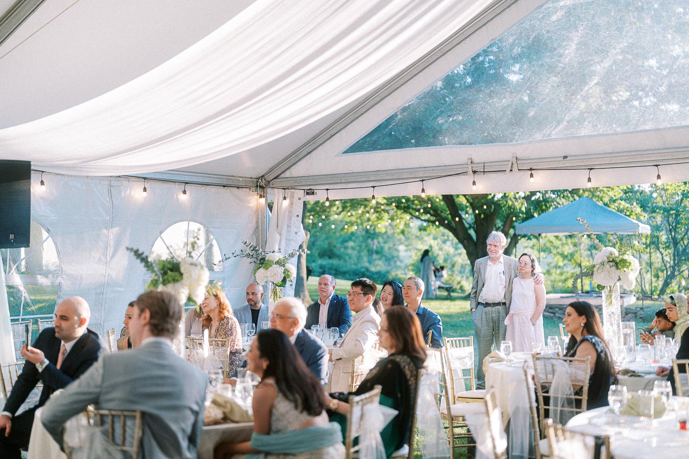 Guests at an elegant outdoor wedding reception sit under a decorated white tent, with natural greenery and floral arrangements enhancing the scenic backdrop.