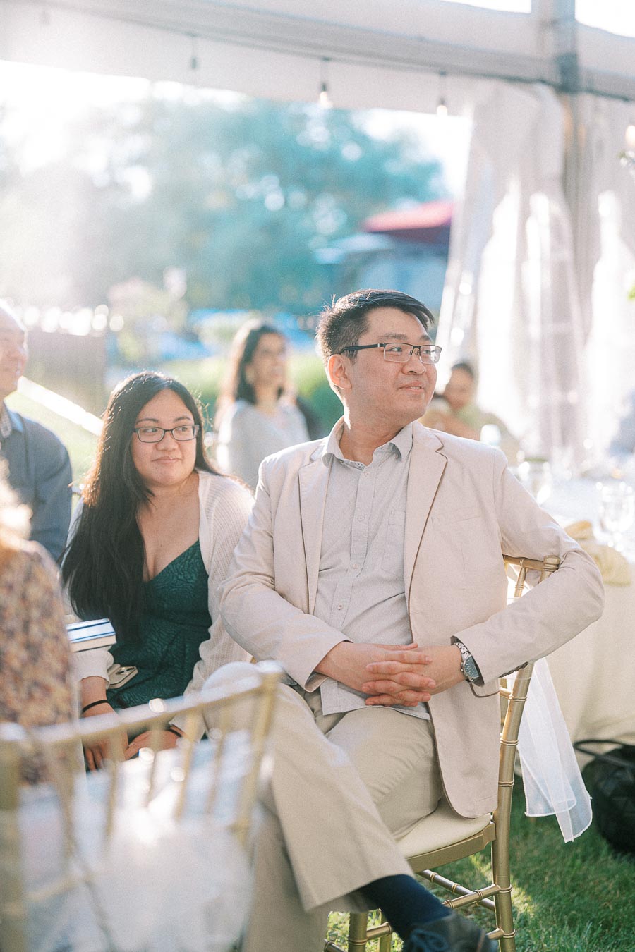 A couple seated at an outdoor event, dressed in formal attire, with natural light streaming in, creating a warm and inviting atmosphere.