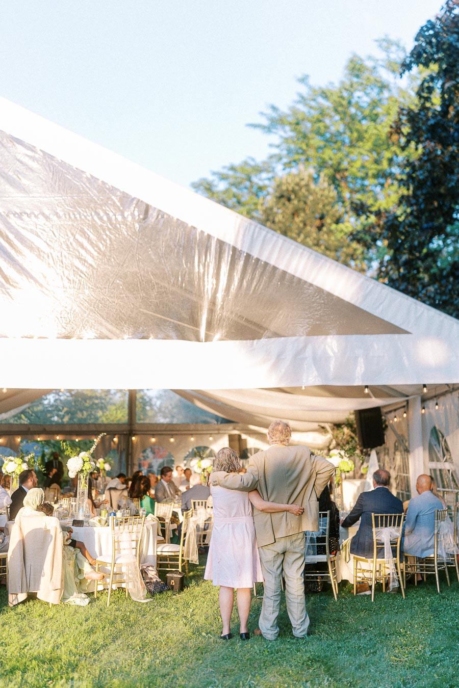 Outdoor wedding reception under a large white tent with guests seated at round tables adorned with floral centerpieces, and a couple embracing in the foreground, enjoying the festive atmosphere on a sunny day.
