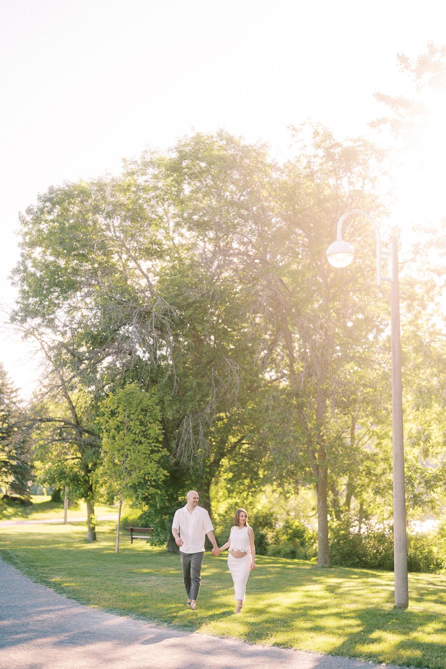 Expectant couple holding hands while walking through a sunny park near Scarborough Bluffs.