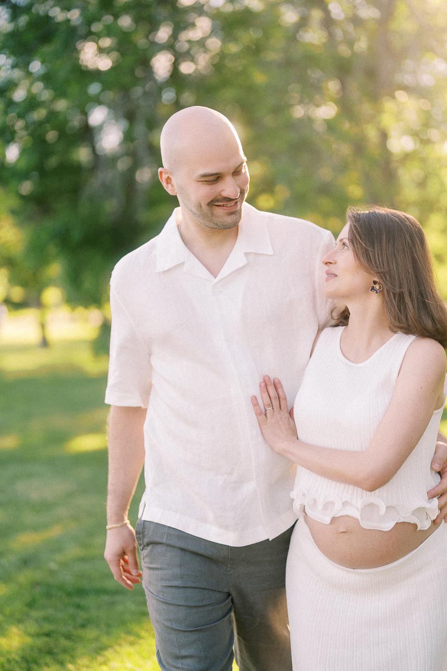 Expectant couple in white outfits walking together through a sunlit green park.