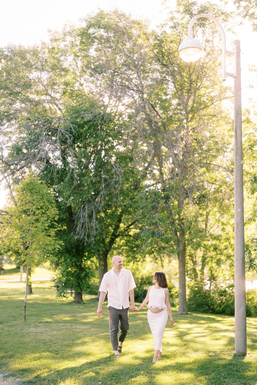 Expectant couple holding hands while walking on grass beneath trees and a lamp post.