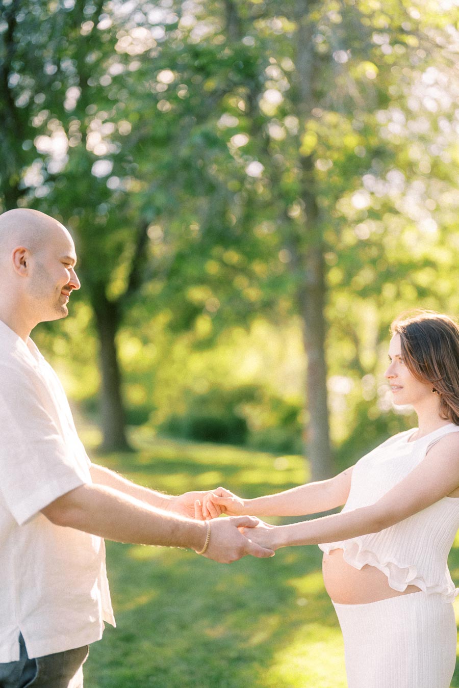 Pregnant woman and partner holding hands in a sunlit green park.