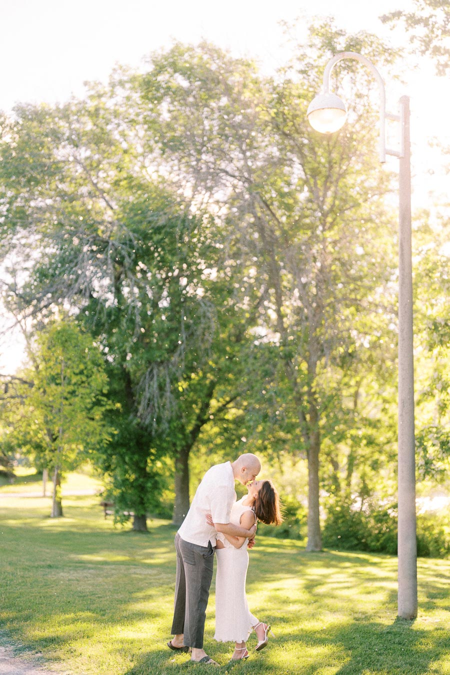 Expectant couple kissing on a sunlit grassy path with trees and a lamp post nearby.