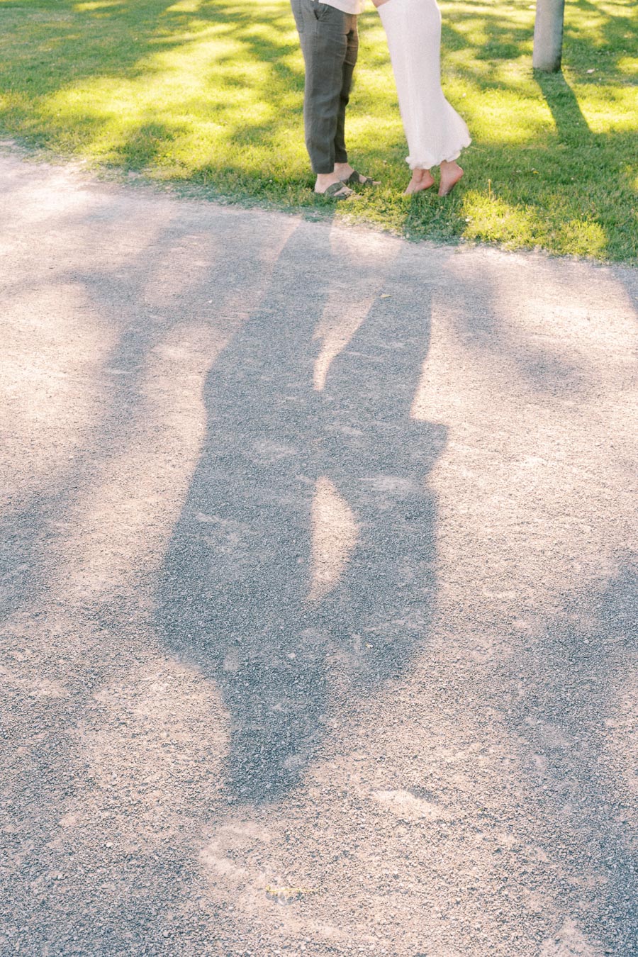 Couple’s lower legs and shadows fall across a sunlit path beside grass.