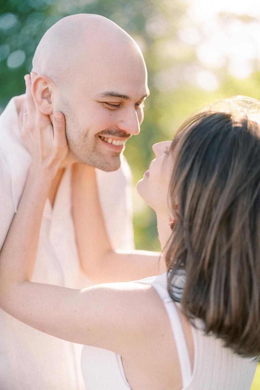 Pregnant woman gently holds her smiling partner’s face in warm outdoor sunlight.