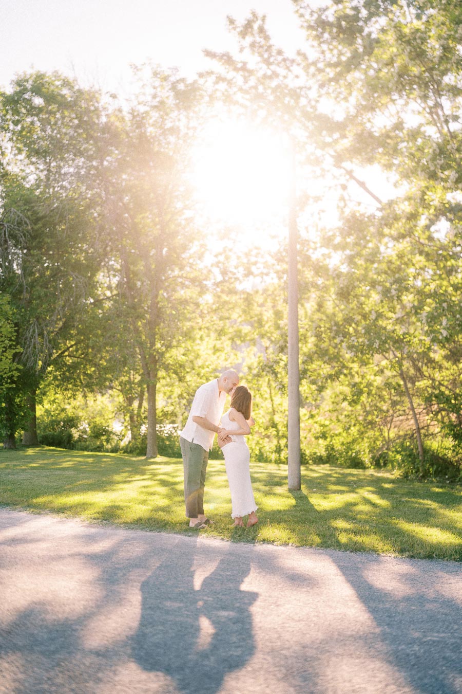Expectant couple kissing on a sunlit park path surrounded by trees.