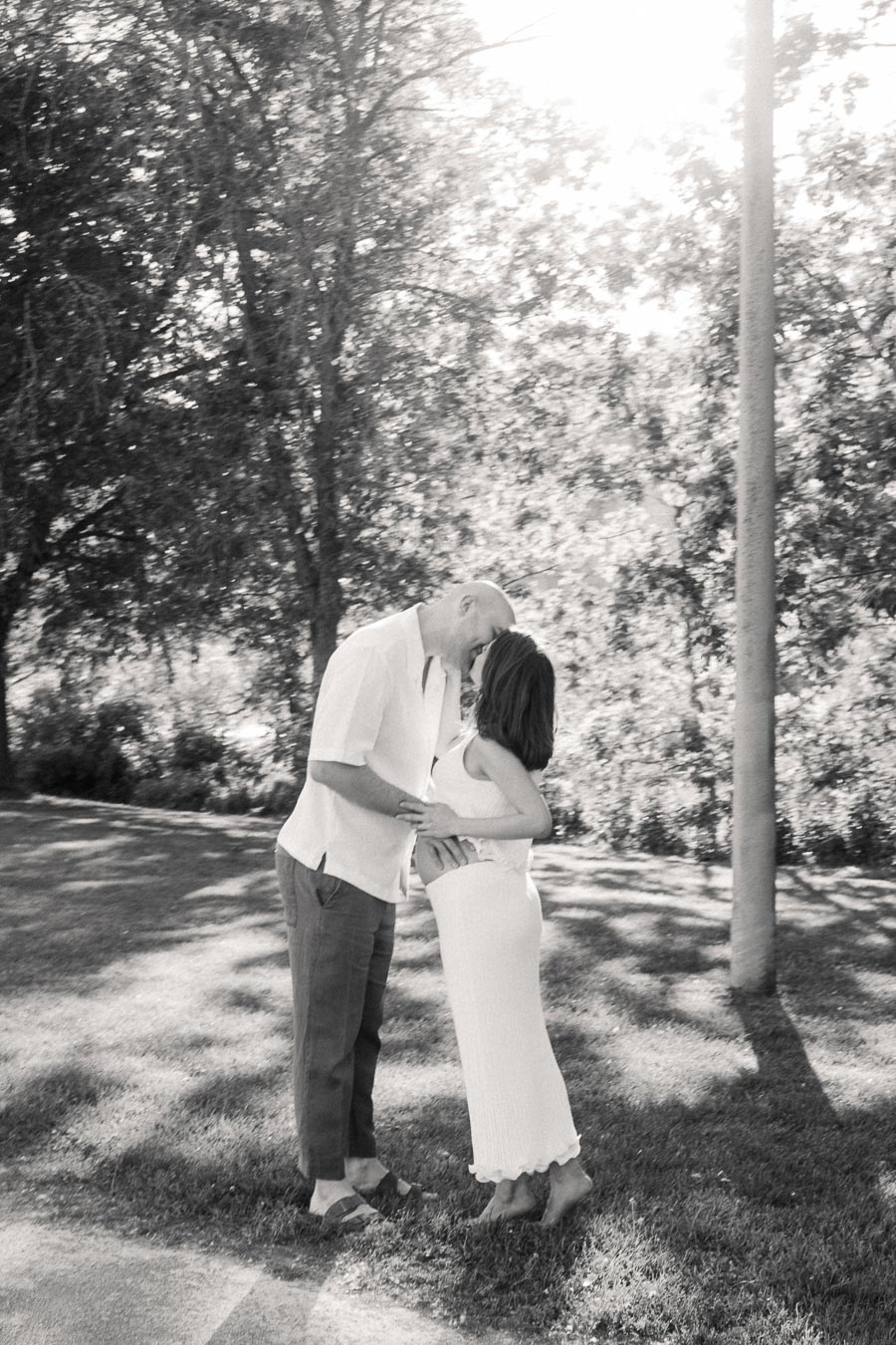 Expectant couple standing forehead to forehead on grass beneath trees in black and white.