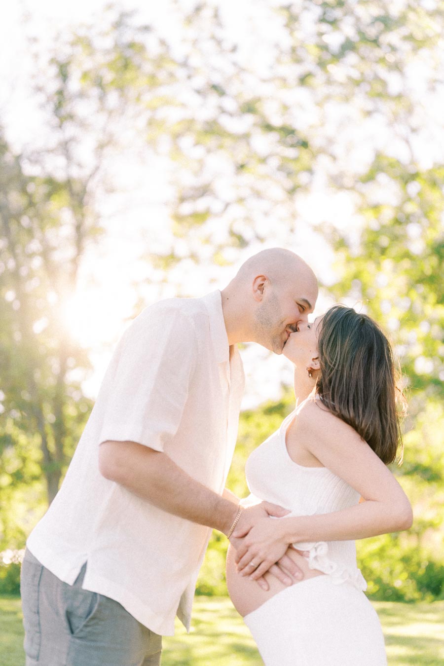 Pregnant couple kissing while holding her belly in soft sunlight among green trees.