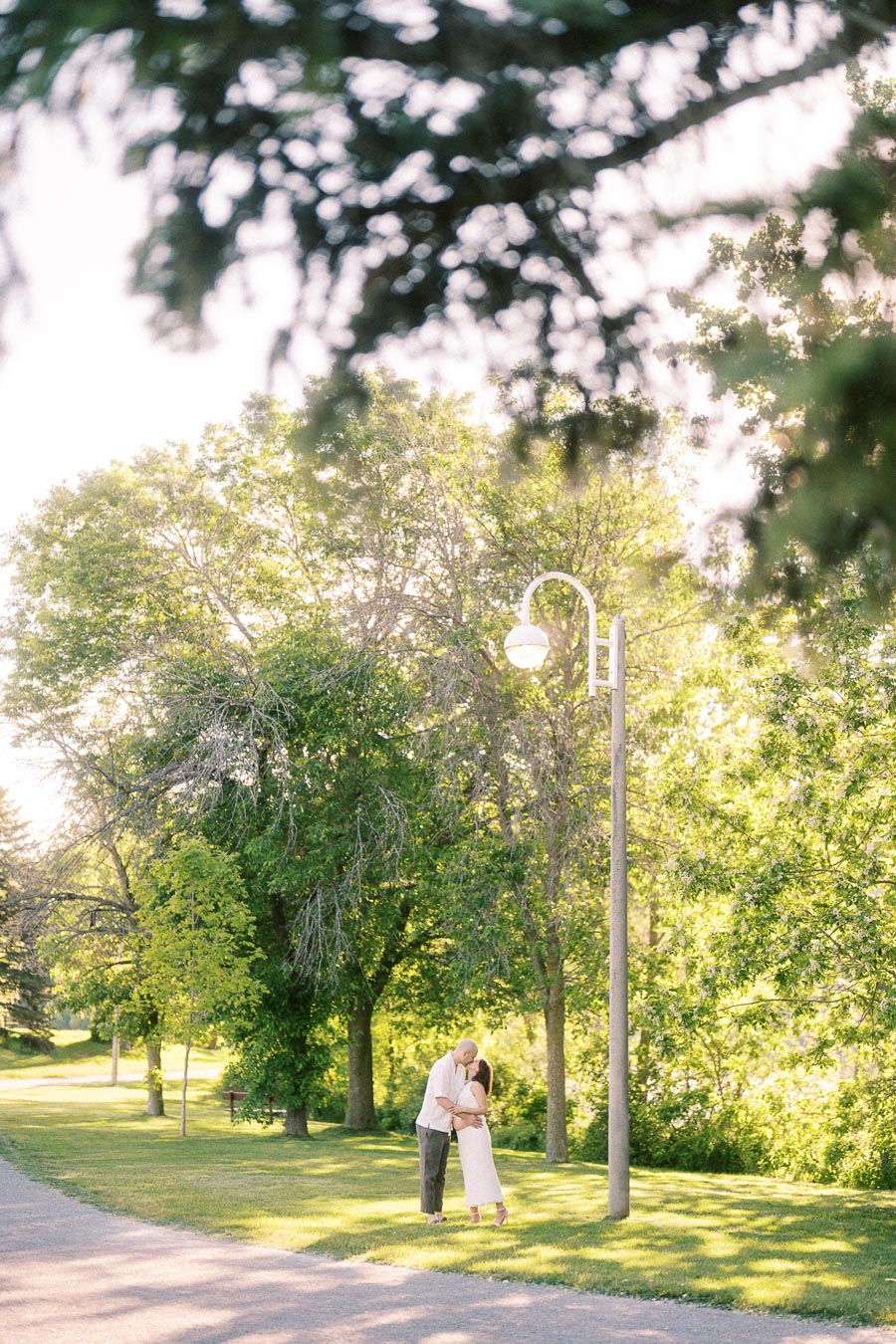 Expectant couple kissing under trees beside a path and lamppost in a sunny park.
