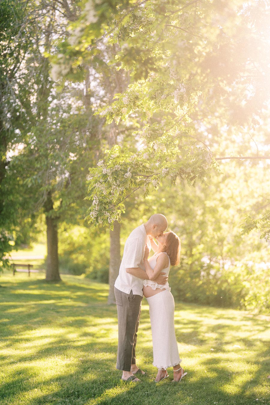 Expectant couple kissing under sunlit trees in a grassy park at Scarborough Bluffs.