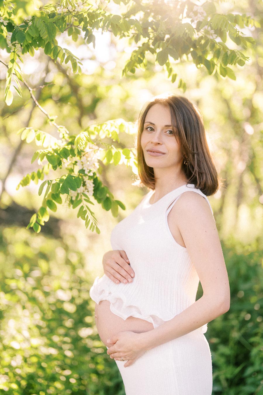 Pregnant woman in a white dress holds her belly under sunlit green branches.