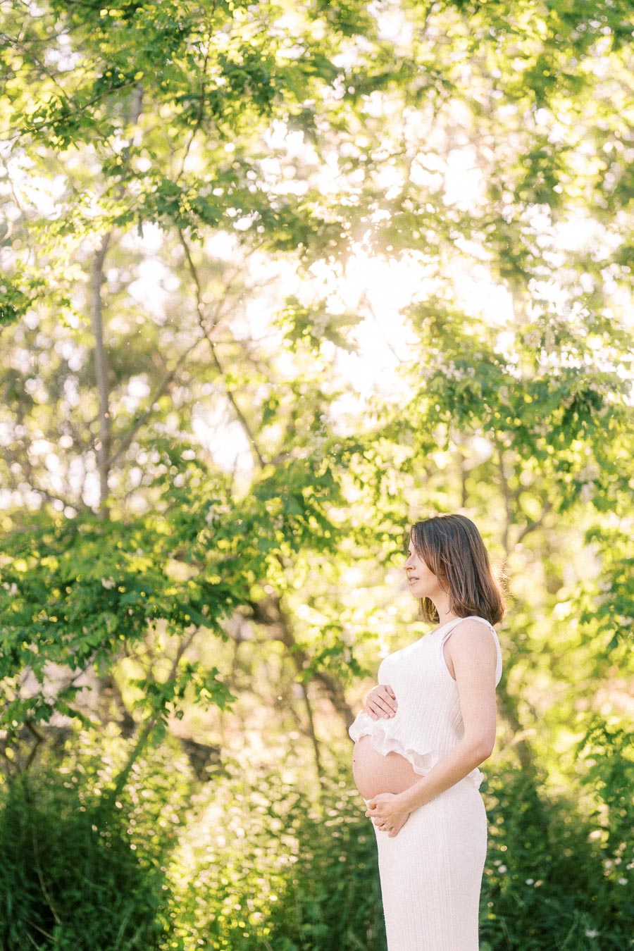 Pregnant woman in a white dress holding her belly beneath sunlit green trees.
