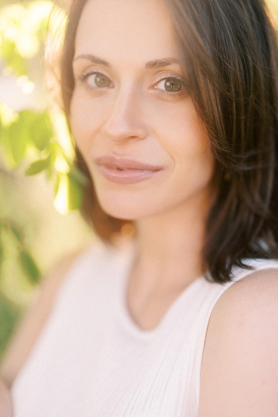 Close-up portrait of a woman in a white sleeveless top with warm sunlight and greenery behind her.