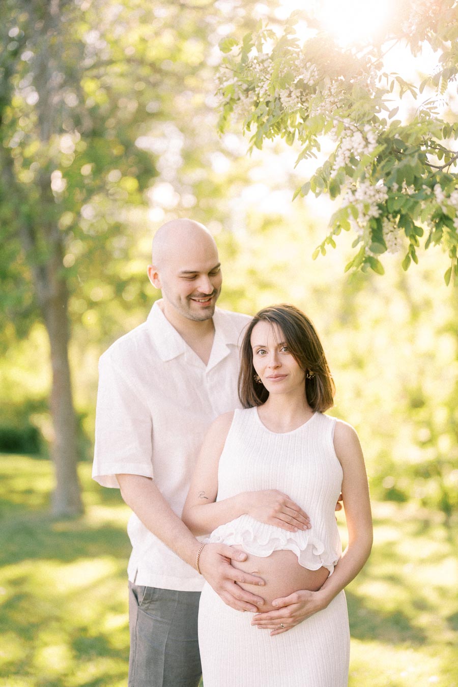Expectant couple in white clothing holding her baby bump under flowering branches in warm sunlight.