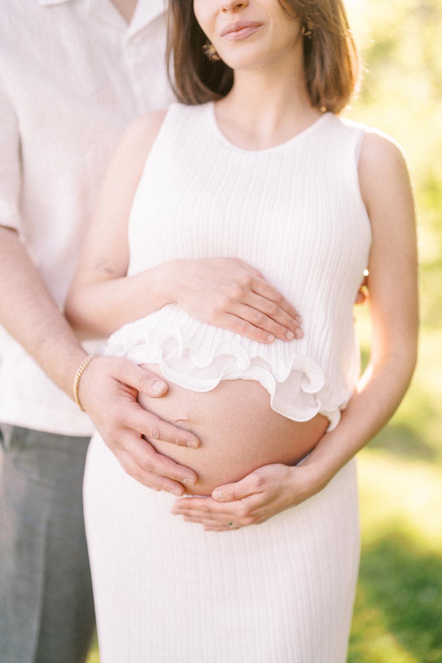 Pregnant woman in a white outfit with her partner’s hands resting on her belly outdoors.