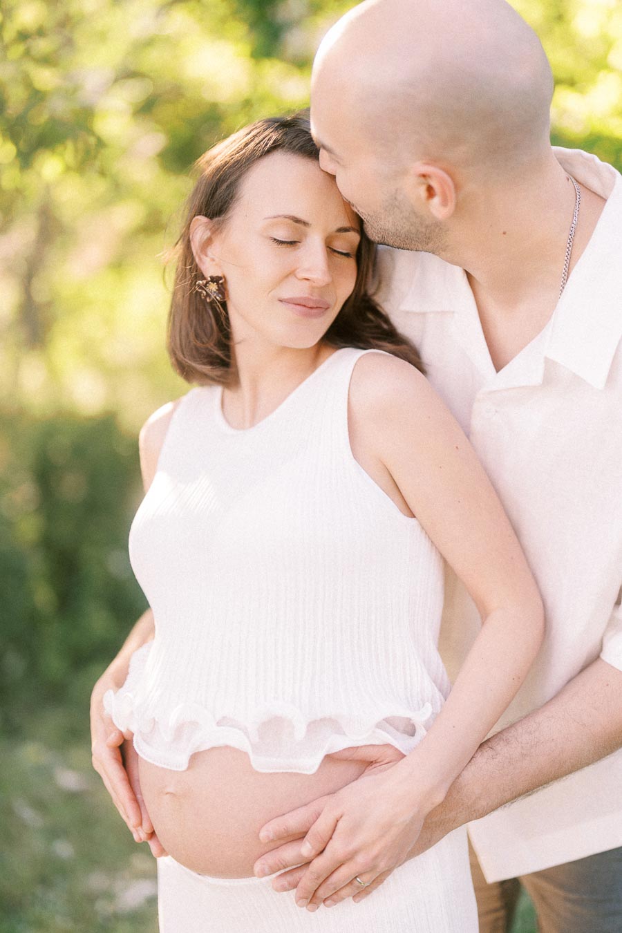 Expectant couple in white clothing embracing while the man kisses the woman’s forehead outdoors.