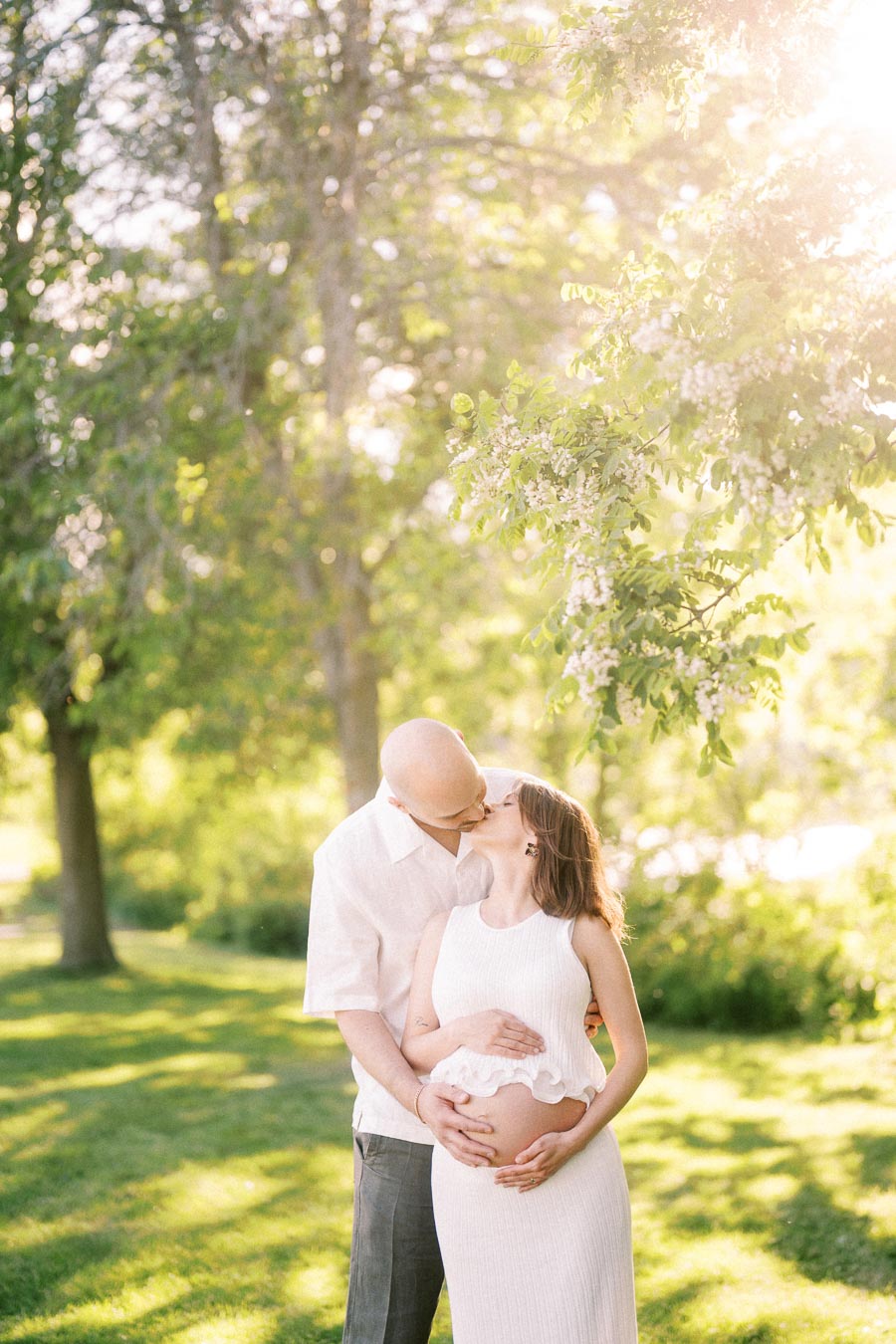 Expectant couple kisses under sunlit trees while holding her pregnant belly.