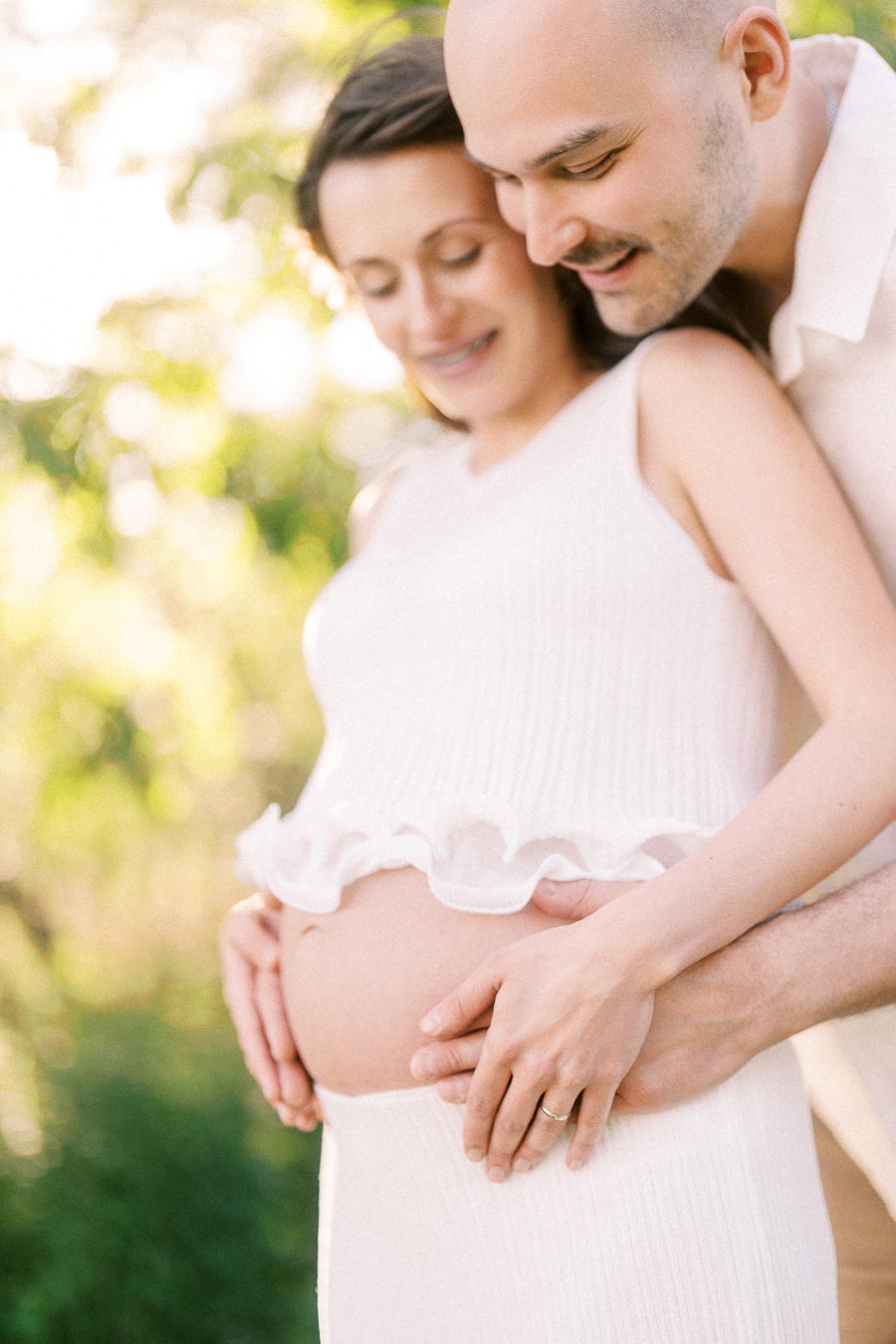 Expectant couple gently holds her baby bump in soft outdoor sunlight.