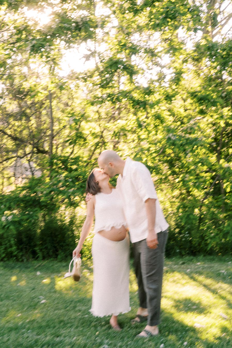Expectant couple kissing in a sunny green park during a maternity portrait session.