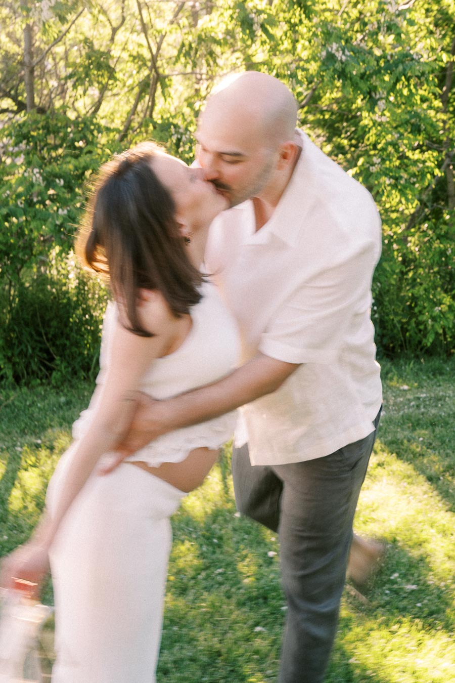 Expectant couple kissing while he dips her in a sunny green park setting.