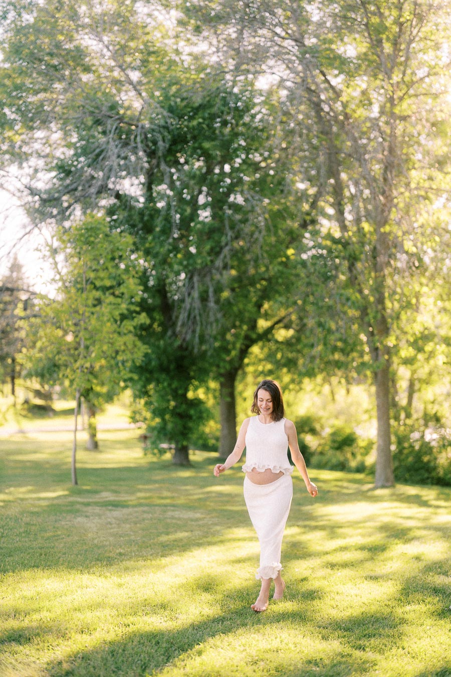 Pregnant woman in white walking barefoot through sunlit grass beneath tall trees.