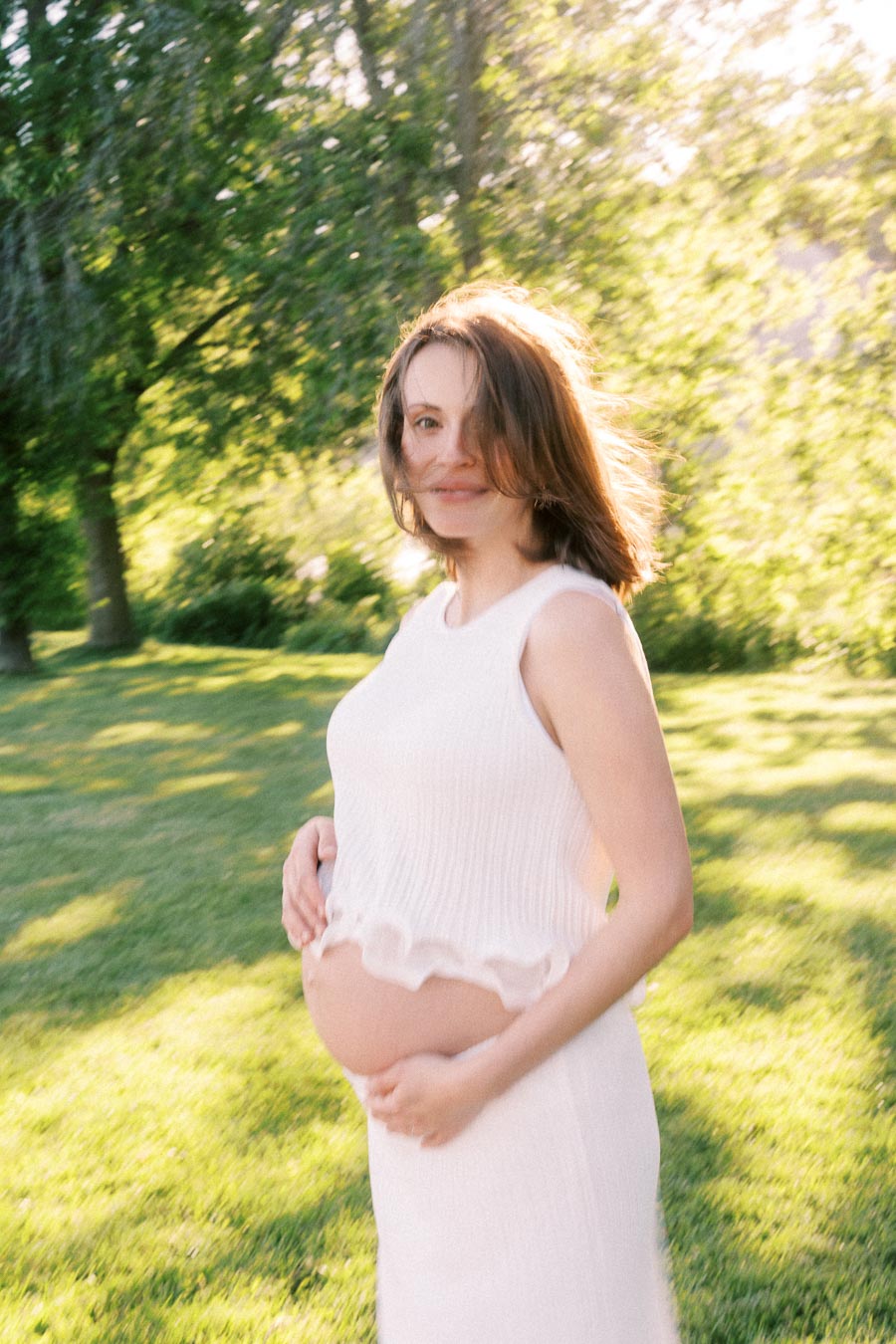 Pregnant woman in a white dress holds her belly in a sunlit green park.