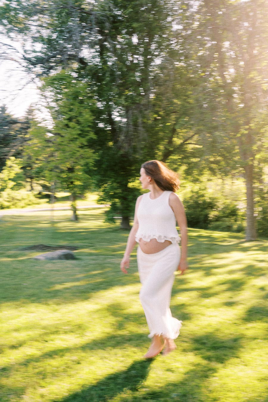 Pregnant woman in a white outfit walking through a sunlit grassy park.