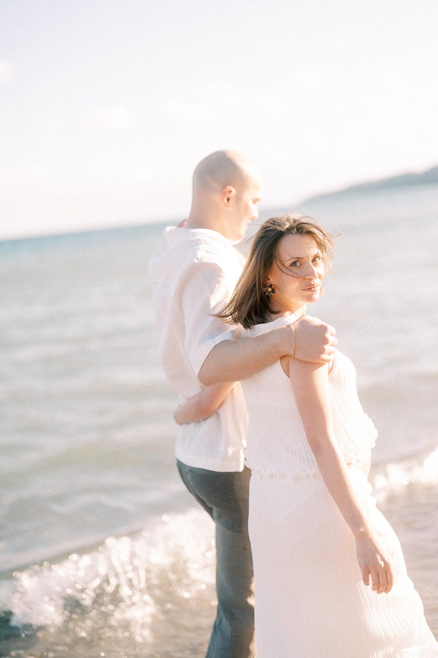 Pregnant woman in white looks back while her partner holds her near the lake.