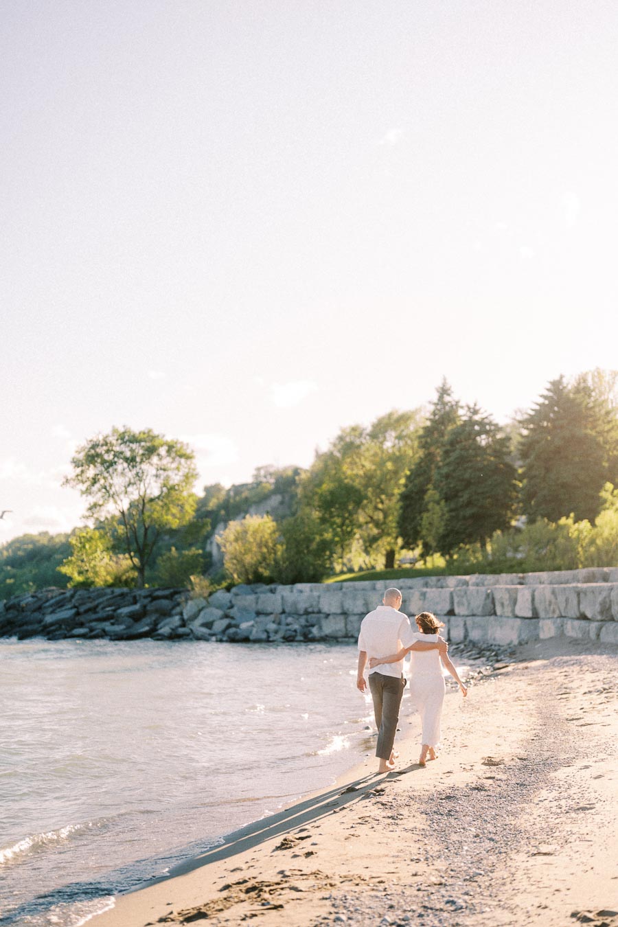 Couple walks arm in arm along a sunny beach beside Lake Ontario.