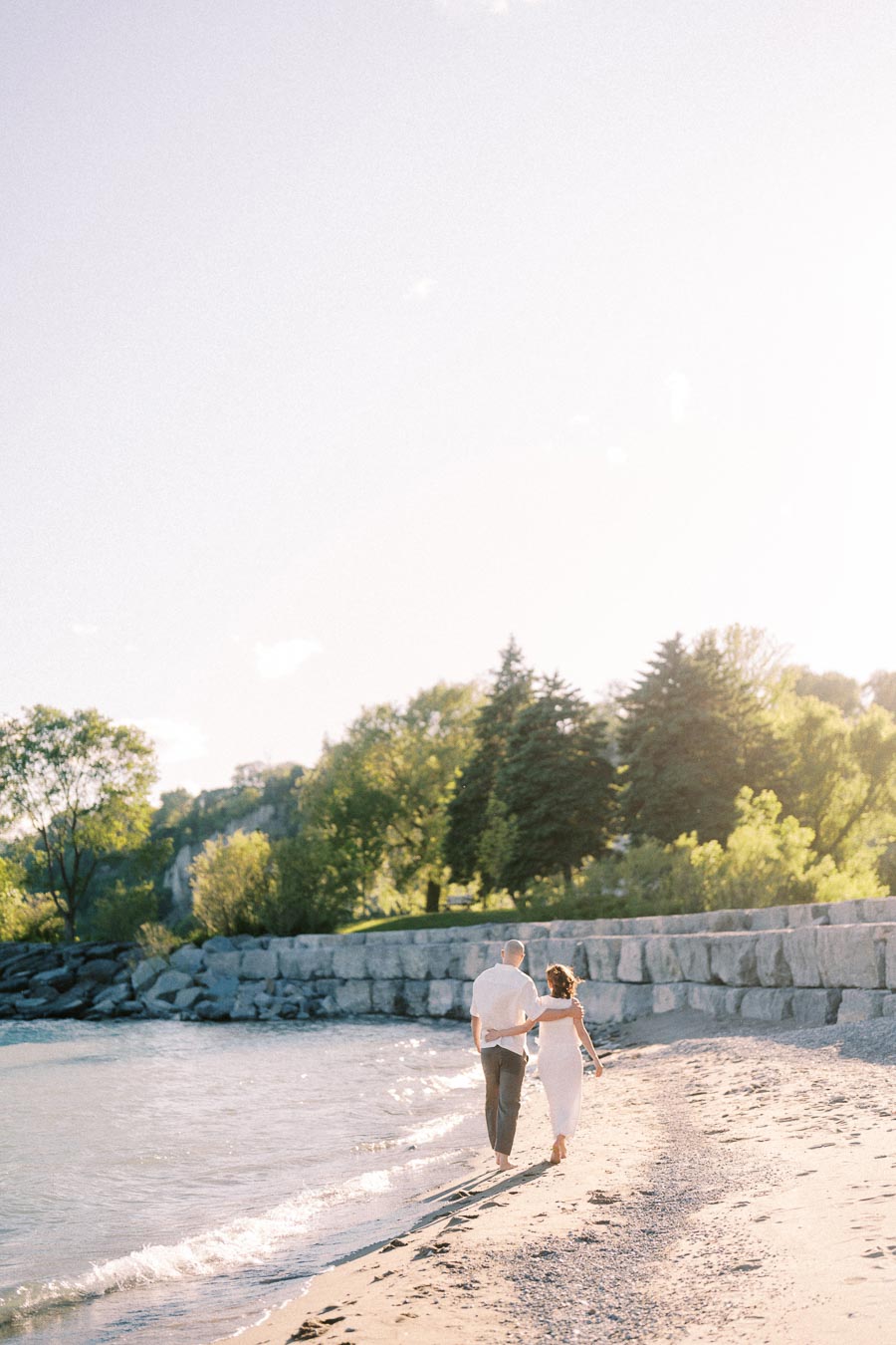 Expectant couple walks arm in arm along a sunny beach beside Lake Ontario.