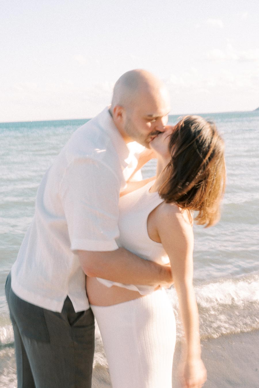 Expectant couple kissing at the shoreline with Lake Ontario waves behind them.