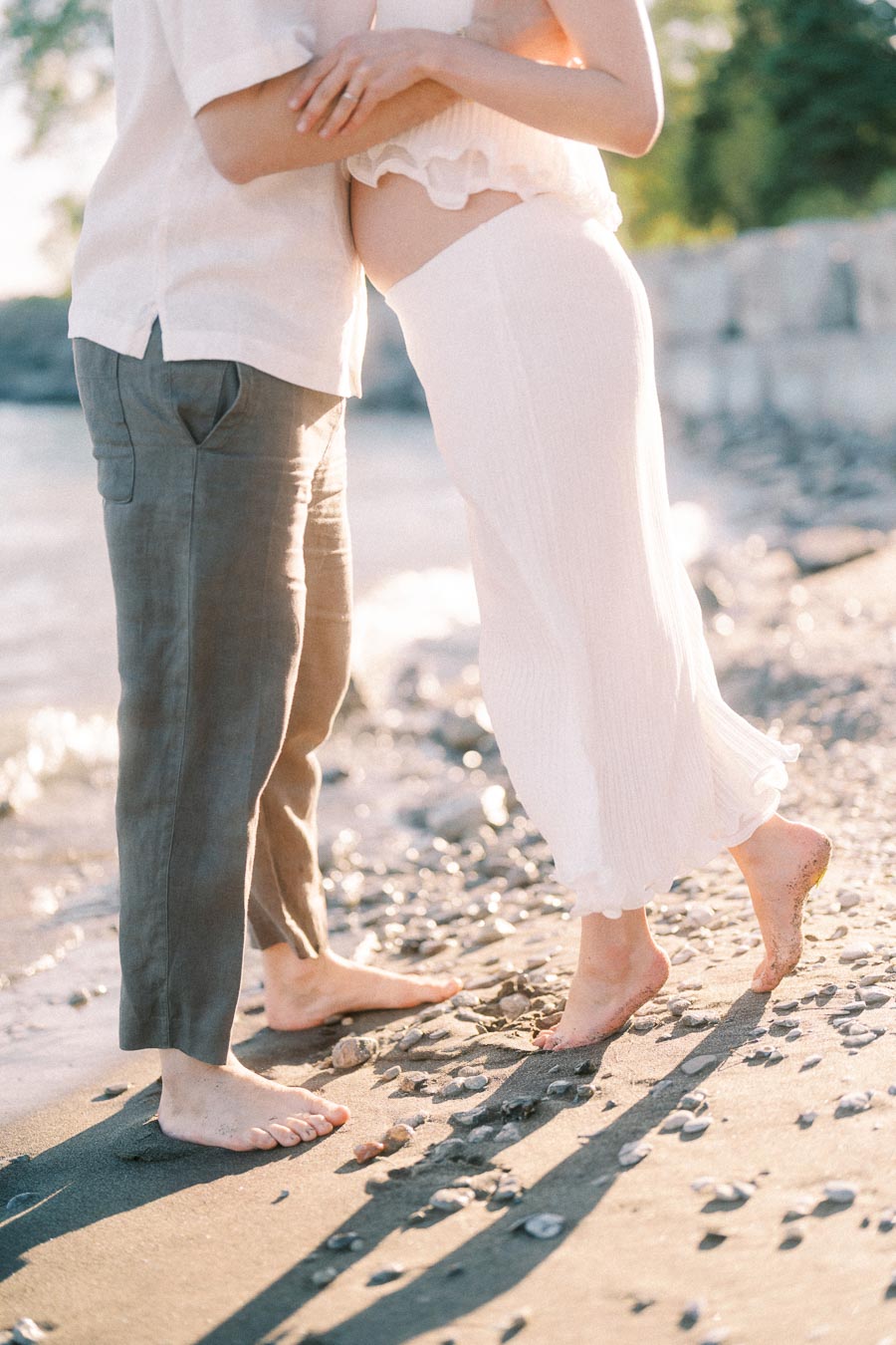 Pregnant couple standing barefoot together on a sunlit pebble beach by the water.