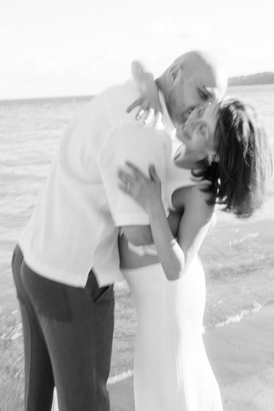 Black-and-white image of a couple embracing and kissing by the water.