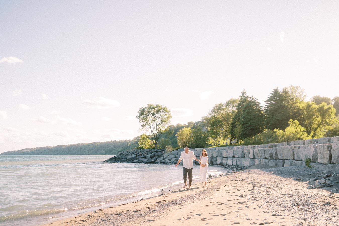 Expectant couple walks hand in hand along a sandy Lake Ontario shoreline.