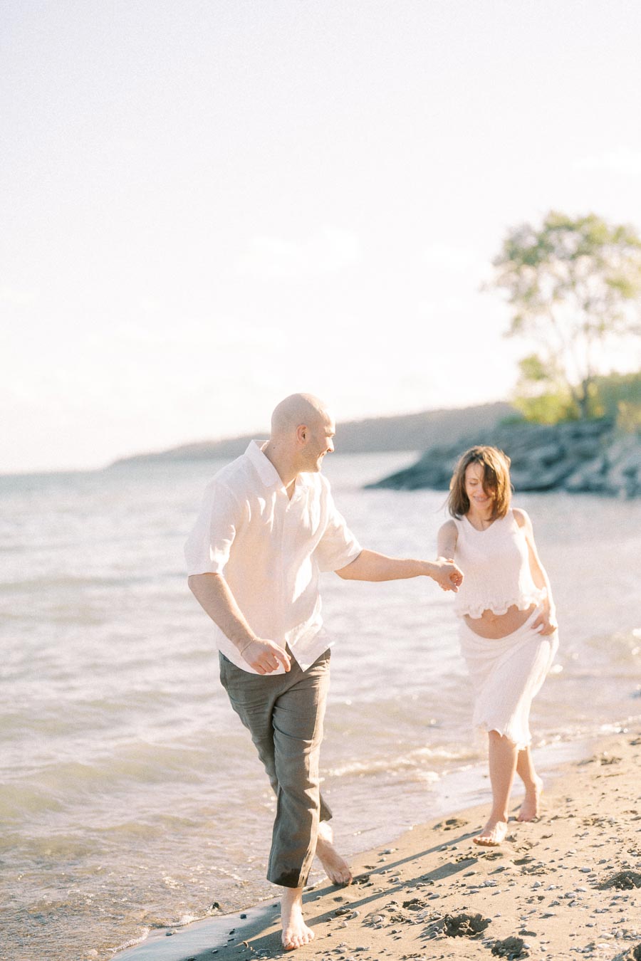 Expectant couple walking barefoot hand in hand along the sandy Lake Ontario shoreline.