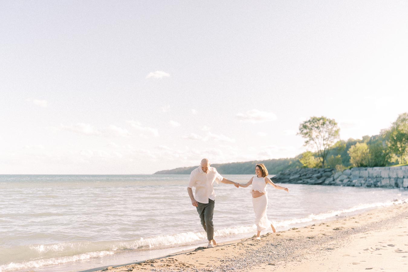Expectant couple holds hands while walking along the beach beside Lake Ontario.