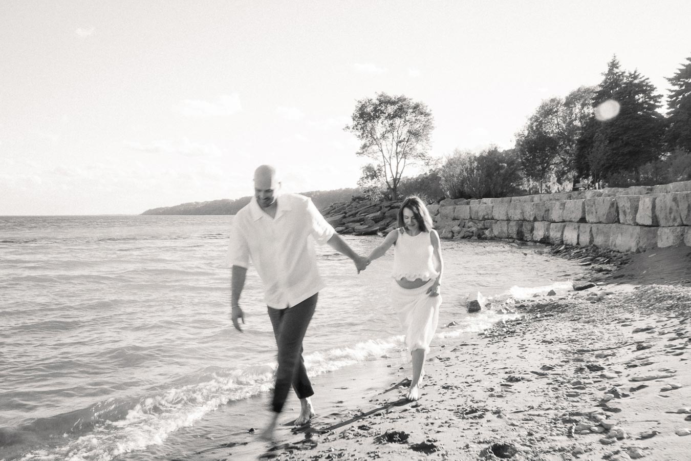 Expectant couple holding hands while walking along the Lake Ontario shoreline.