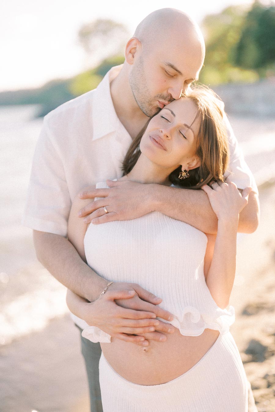 Expectant couple embracing on a sunlit beach, hands resting on the mother's baby bump.