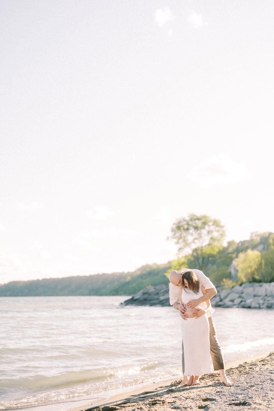 Expectant couple embraces on a sandy Lake Ontario beach at Scarborough Bluffs.