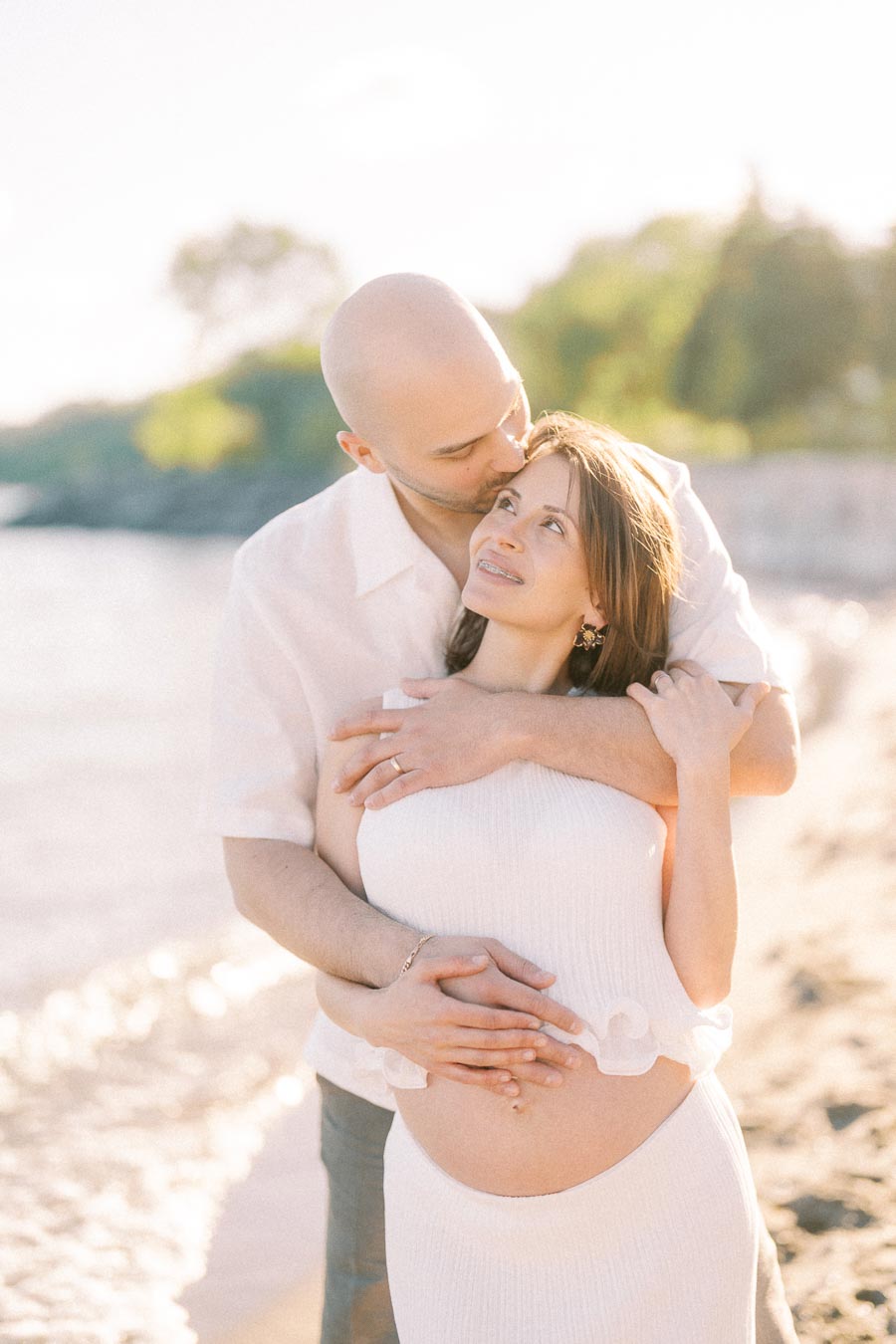 Expectant couple embracing on a sunny beach, with the man kissing the woman's forehead.