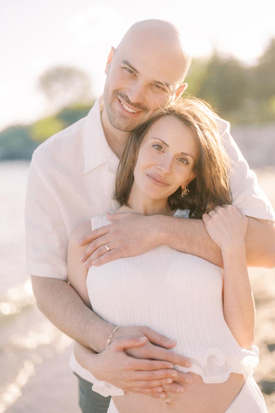 Expectant couple embracing on a sunny beach with hands resting on the pregnant belly.