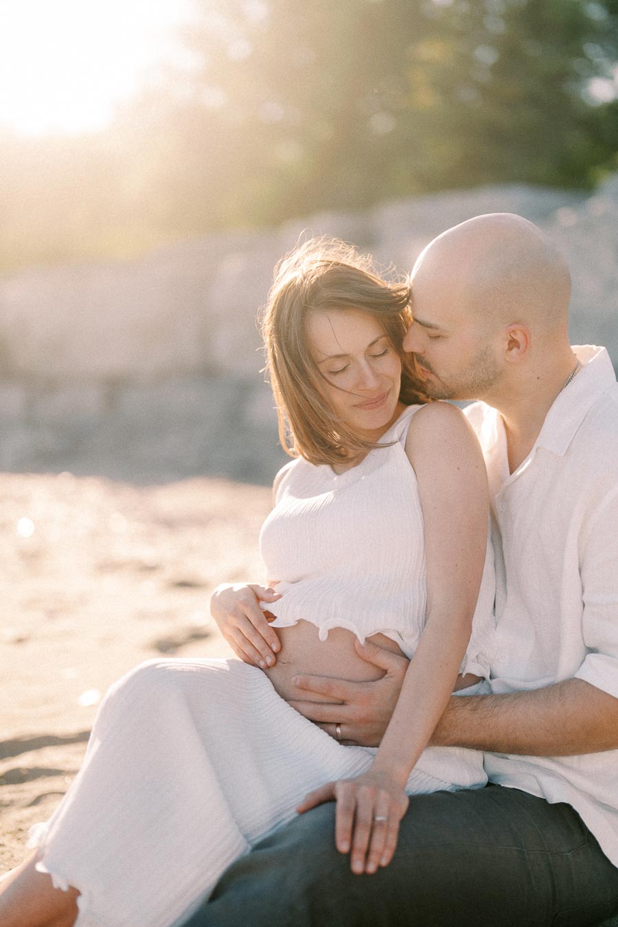 Expectant couple sitting on a sunny beach as the man kisses the woman’s shoulder.
