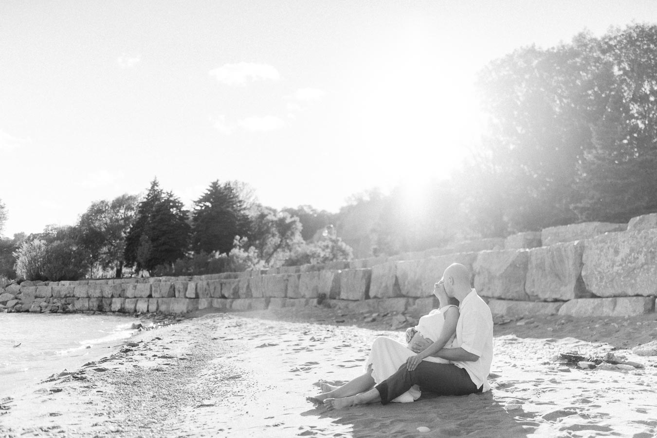 Expectant couple sitting together on a sandy beach beside stone bluffs in bright sunlight.