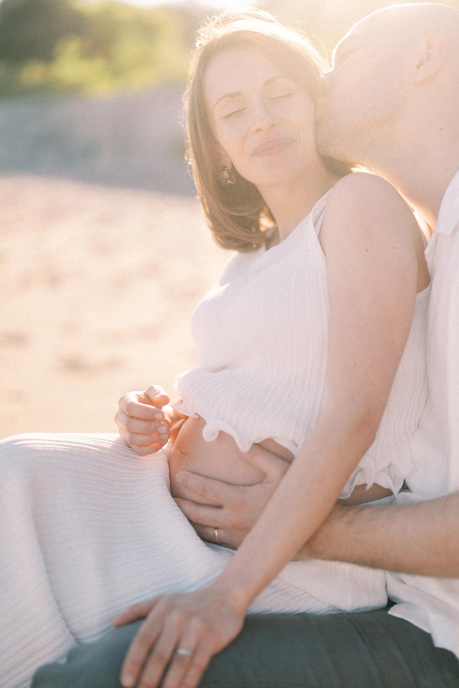 Expectant couple in white clothing cuddles closely as the partner kisses her cheek in warm sunlight.