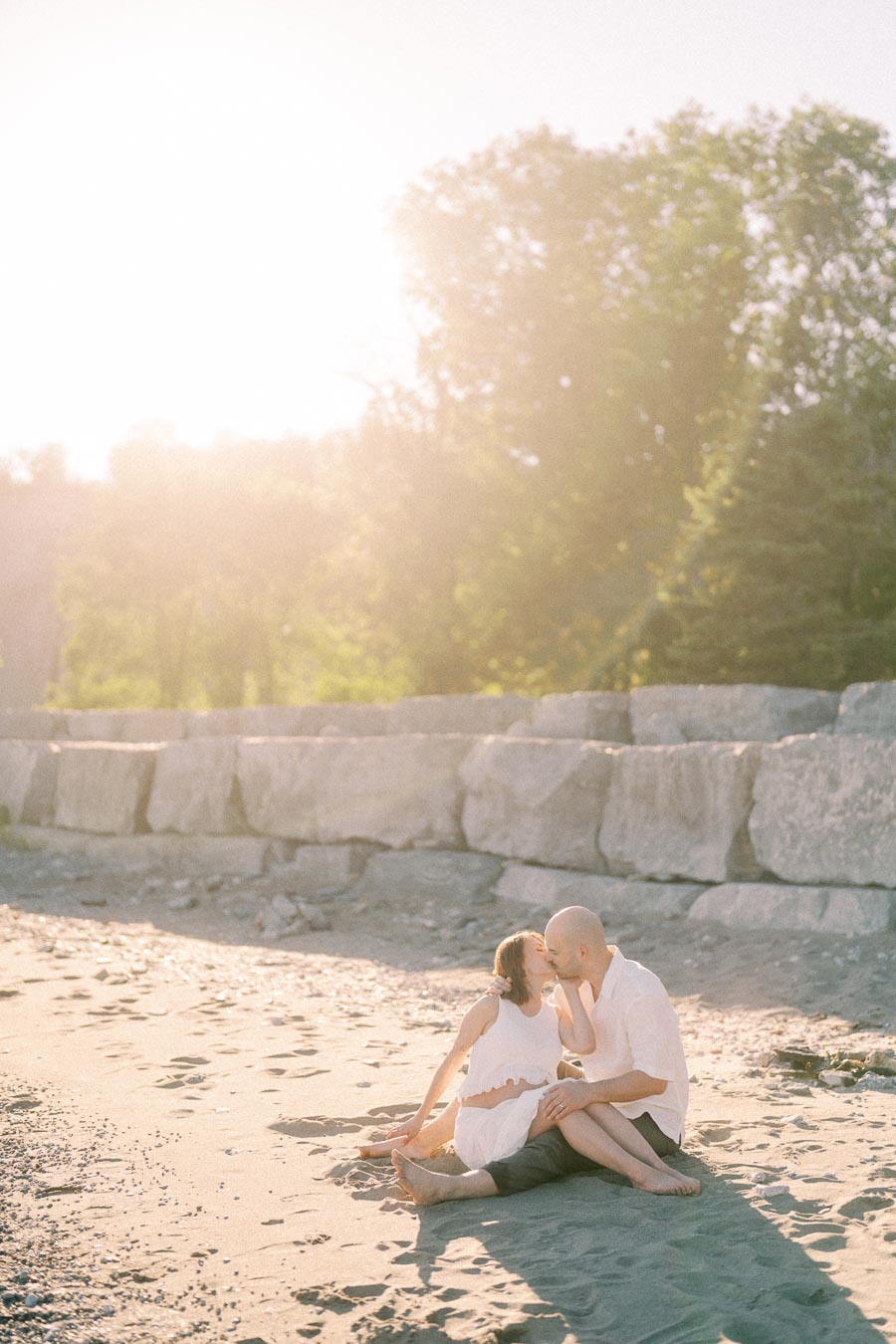 Expectant couple sitting on a sandy beach and kissing in warm evening light.