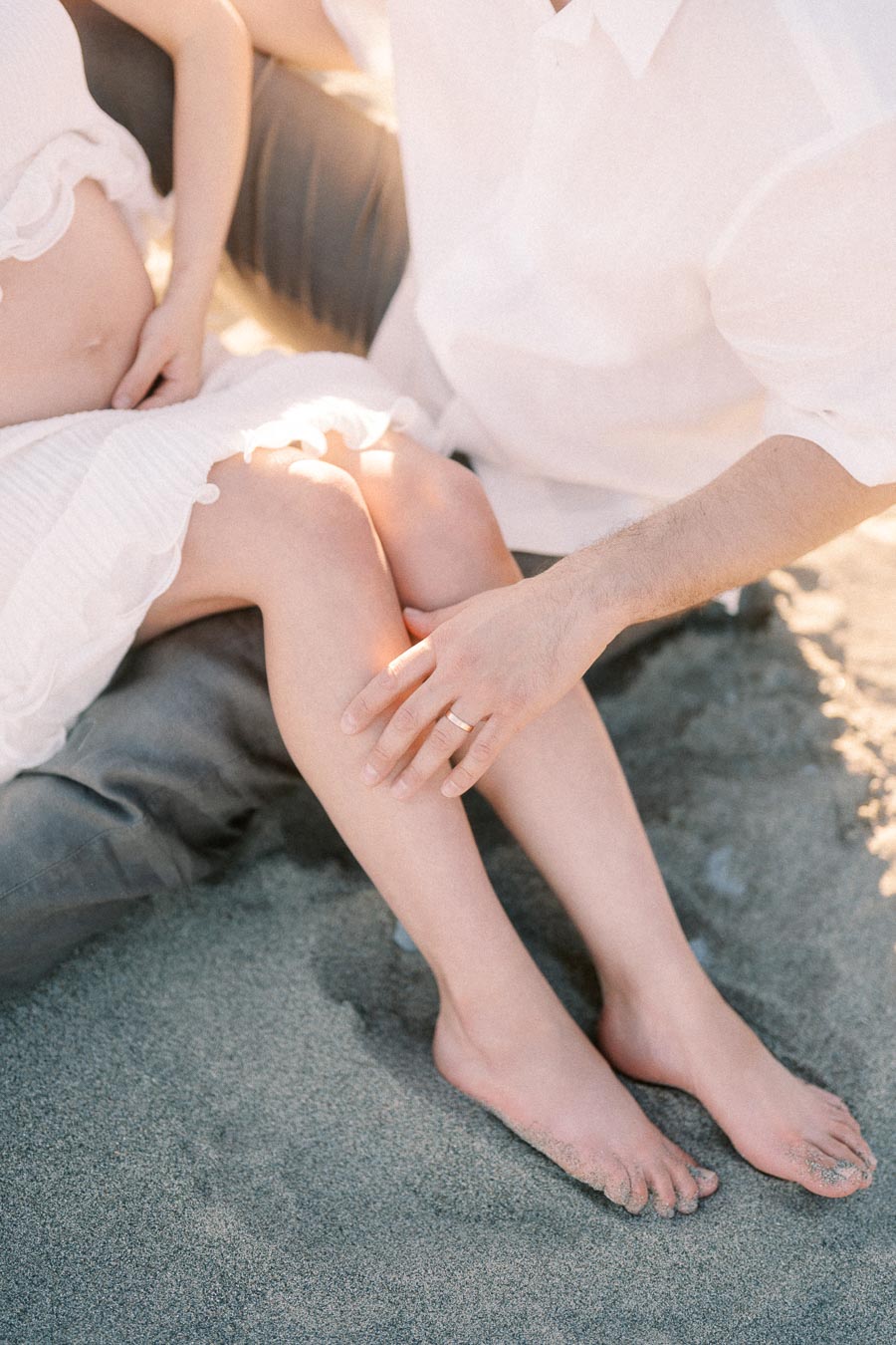 Close-up of a pregnant couple sitting barefoot on sand, with the father’s hand on her leg.
