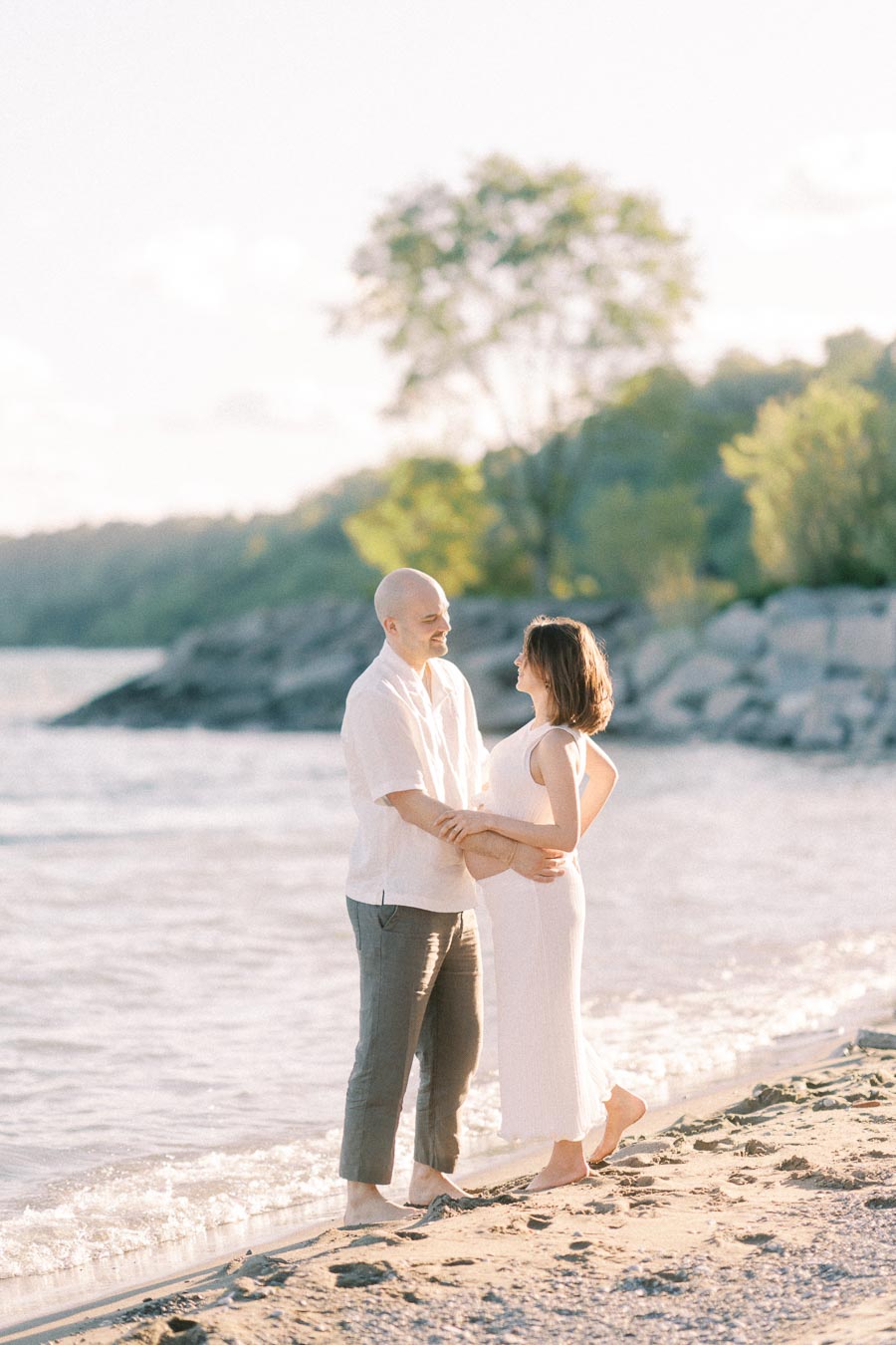 Expectant couple holding hands on a sandy beach beside Lake Ontario in warm evening light.